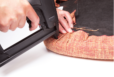 Mains utilisant une agrafeuse pour fixer un tissu orange sur une assise.